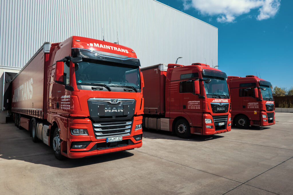 Drei rote LKW von Maintrans stehen an der Laderampe eines Logistik-Lagers und werden beladen.