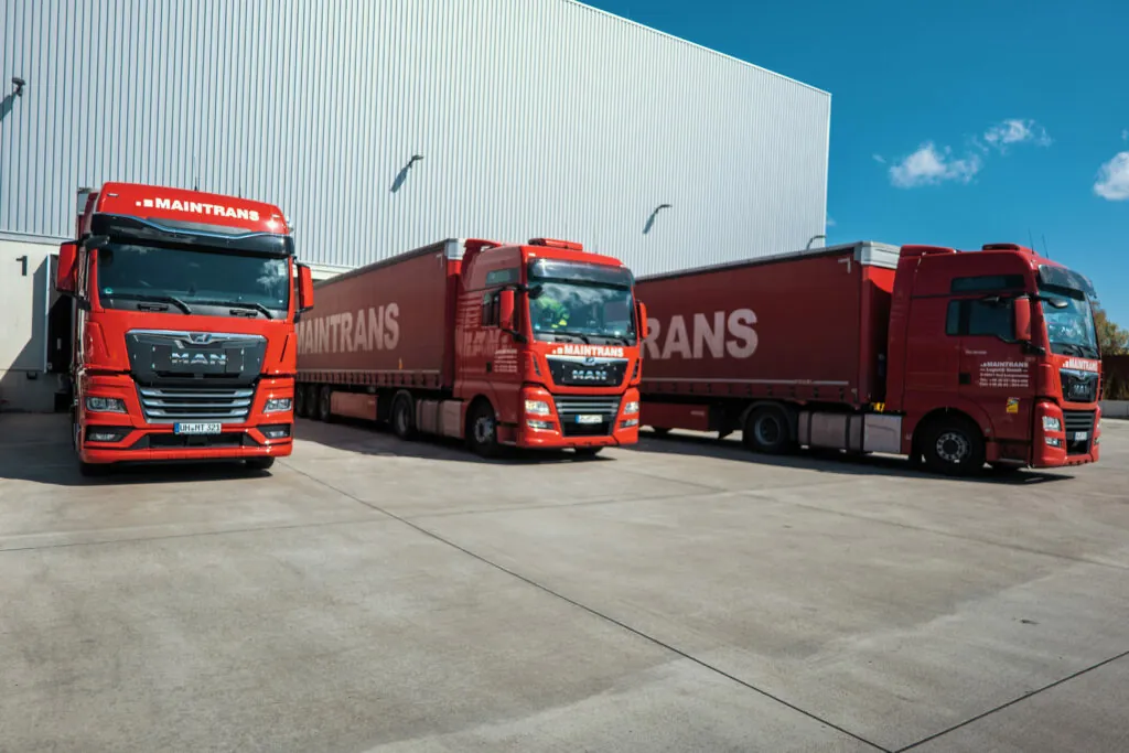 Drei rote LKW mit Maintrans Logo stehen nebeneinander an der Laderampe eines Logistik-Lagers.