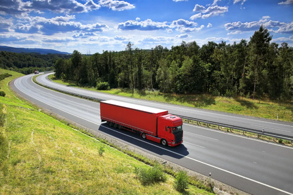 Ein roter Transport-LKW fährt auf einer Autobahn, die durch eine grüne, bewaldete Landschaft führt.